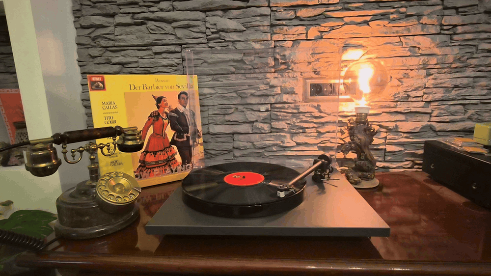 A turntable with a spinning record, beside a vintage telephone and a lamp against a stone wall