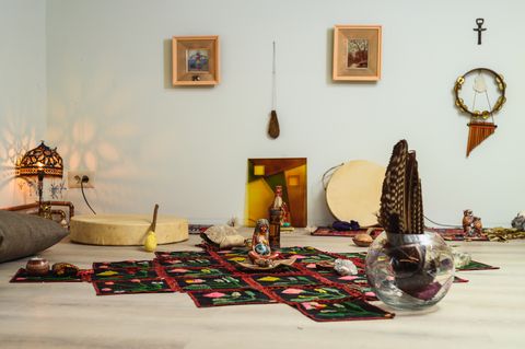 Embroidered rugs on the floor, a goddess figure, a glass vessel with feathers and a drum by the wall